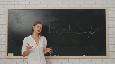 Okul öğretmeni sınıfta öğrencilere konuşuyor. Sınıftaki genç bayan üniversite öğretmeni öğrencilere İngilizce dersini karatahtanın önünde açıklıyor. Eğitim kavramı.