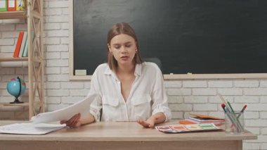 Okul öğretmeni sınıfta öğrencilere konuşuyor. Tahtanın önündeki masada oturan genç bayan üniversite öğretmeni ödevlerini öğrencilere kızgın bir şekilde anlatıyor. Eğitim kavramı.