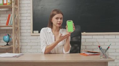 Okul öğretmeni sınıfta çalışır. Sınıftaki masada oturan genç bayan üniversite öğretmeni akıllı telefon kromu yeşil ekran tutuyor ve baş parmağını gösteriyor..