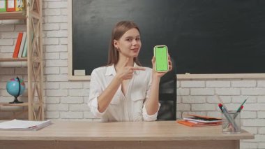 Okul öğretmeni sınıfta çalışır. Sınıftaki masada oturan genç bayan üniversite öğretmeni akıllı telefon kromu yeşil ekran tutuyor ve baş parmağını gösteriyor..