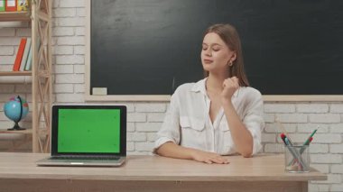 Okul öğretmeni sınıfta öğrencilere konuşuyor. Sınıfın önündeki masada oturan genç bayan kolej öğretmeni öğrencilere dersi açıklıyor, dizüstü bilgisayar krom anahtar yeşil ekran. Eğitim kavramı.