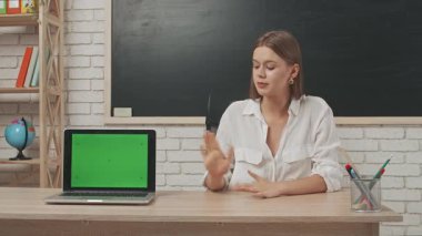 Okul öğretmeni sınıfta öğrencilere konuşuyor. Sınıfın önündeki masada oturan genç bayan kolej öğretmeni öğrencilere dersi açıklıyor, dizüstü bilgisayar krom anahtar yeşil ekran. Eğitim kavramı.