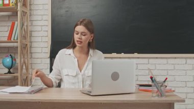 Okul öğretmeni sınıfta öğrencilere konuşuyor. Sınıfın masasındaki yazı tahtasının önünde oturan genç bayan üniversite öğretmeni öğrencilere laptoptaki video çağrısından ders veriyor. Eğitim kavramı.