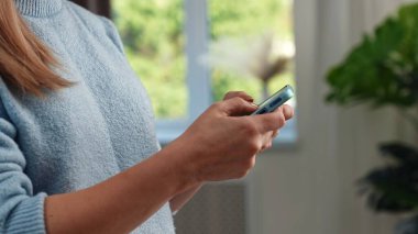 Mavi kazaklı bir kadın, akıllı telefonunu yeşil manzaralı bir pencerenin yanında kullanıyor..