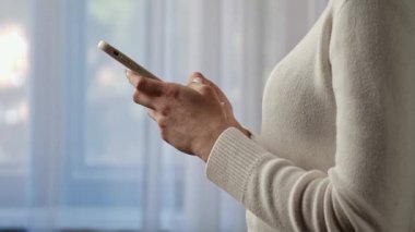 Beyaz kazaklı bir kadın akıllı telefonunu pencerenin yanında kullanıyor ve cihaza bağlanıyor. Yavaş çekim