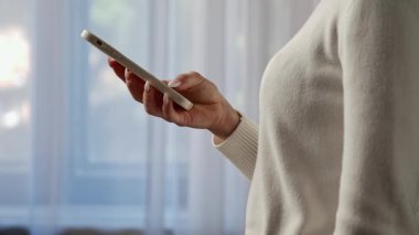Beyaz kazaklı bir kadın akıllı telefonunu pencerenin yanında kullanıyor ve cihaza bağlanıyor. Yavaş çekim