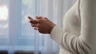 Beyaz kazaklı bir kadın akıllı telefonunu pencerenin yanında kullanıyor ve cihaza bağlanıyor. Yavaş çekim