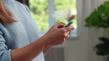 Mavi kazaklı bir kadın, akıllı telefonunu yeşil manzaralı bir pencerenin yanında kullanıyor ve cihaza bağlanıyor. Yavaş çekim