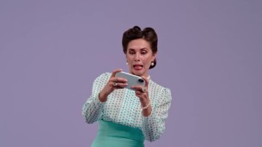 Mor stüdyo arka planında çekici saç stili pozları olan klasik kıyafetli genç bir kadın. Telefonunda retro tarzı oyun oynayan güzel bir aktris ve kazanmaktan mutlu. Güzellik ve eski tarz konsept