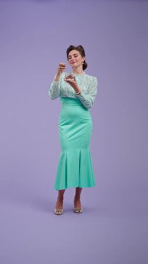 Mor stüdyo arka planında çekici saç stili pozları olan klasik kıyafetli genç bir kadın. Geçmişe dönük güzel bir aktris parfümünü zevkle üzerine sıkıyor. Güzellik ve eski tarz konsept.