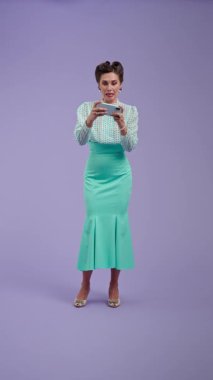 Mor stüdyo arka planında çekici saç stili pozları olan klasik kıyafetli genç bir kadın. Telefonunda retro tarzı oyun oynayan güzel bir aktris ve kazanmaktan mutlu. Güzellik ve eski tarz konsept