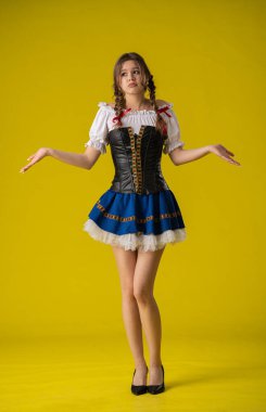 Geleneksel bir kostümle genç bir kadın, parlak sarı bir zemin karşısında şaşkın bir ifadeyle ayakta duruyor..