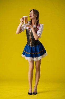 Geleneksel giyinmiş bir kadın, parlak sarı bir arka planda duran büyük bir kupa biranın keyfini çıkarıyor..