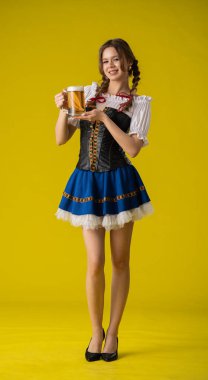 Geleneksel giyinmiş bir kadın, parlak sarı bir arka planda duran büyük bir kupa biranın keyfini çıkarıyor..