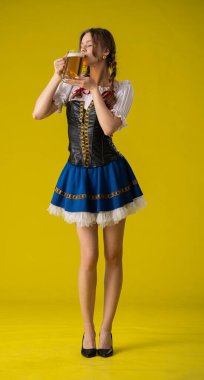 Geleneksel giyinmiş bir kadın, parlak sarı bir arka planda duran büyük bir kupa biranın keyfini çıkarıyor..