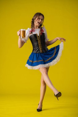 Geleneksel giyinmiş bir kadın, parlak sarı bir arka planda duran büyük bir kupa biranın keyfini çıkarıyor..