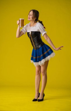 Geleneksel giyinmiş bir kadın, parlak sarı bir arka planda duran büyük bir kupa biranın keyfini çıkarıyor..