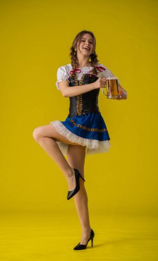 Geleneksel giyinmiş bir kadın, parlak sarı bir arka planda duran büyük bir kupa biranın keyfini çıkarıyor..