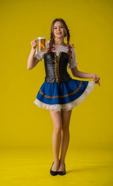 Geleneksel giyinmiş bir kadın, parlak sarı bir arka planda duran büyük bir kupa biranın keyfini çıkarıyor..