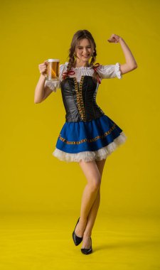 Geleneksel giyinmiş bir kadın, parlak sarı bir arka planda duran büyük bir kupa biranın keyfini çıkarıyor..