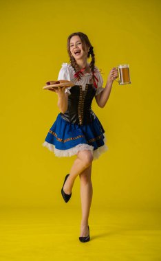 Geleneksel giyinmiş genç bir kadın elinde bir bardak bira ve sosisle sarı arka planda..