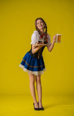 Geleneksel giyinmiş genç bir kadın elinde bir bardak bira ve sosisle sarı arka planda..