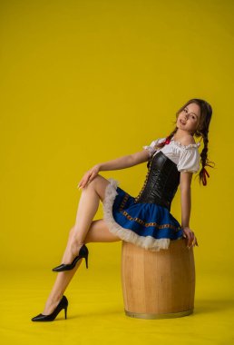 Geleneksel bir kostüm giymiş bir kadın zarif bir şekilde tahta bir fıçının üzerinde oturur. Parlak sarı arka plan onun kıyafetlerini ön plana çıkarıyor..