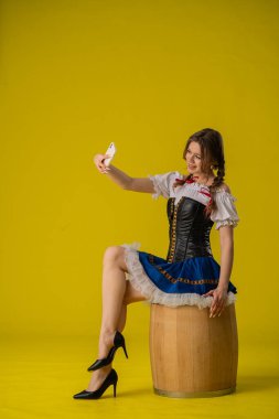 Geleneksel Alman Dirndl elbisesi giymiş genç bir kadın, parlak sarı bir arka plana karşı tahta bir fıçının üzerinde otururken selfie için poz veriyor..