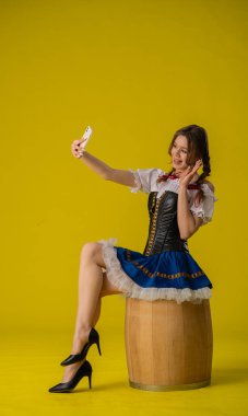 Geleneksel Alman Dirndl elbisesi giymiş genç bir kadın, parlak sarı bir arka plana karşı tahta bir fıçının üzerinde otururken selfie için poz veriyor..