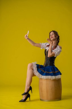Geleneksel Alman Dirndl elbisesi giymiş genç bir kadın, parlak sarı bir arka plana karşı tahta bir fıçının üzerinde otururken selfie için poz veriyor..