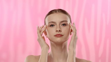 Açık tenli genç bir kadın nazikçe yüzüne dokunuyor, cilt teması ve gençlik ışıltısı öneriyor..