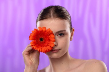 Genç bir kadın parlak turuncu bir gerbera papatyasını gözünün önünde canlı mor bir arka planda tutuyor..