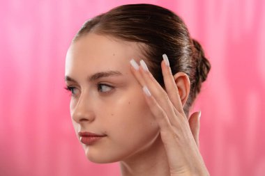 Açık tenli genç bir kadın nazikçe yüzüne dokunuyor, cilt teması ve gençlik ışıltısı öneriyor..