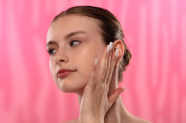 Açık tenli genç bir kadın nazikçe yüzüne dokunuyor, cilt teması ve gençlik ışıltısı öneriyor..