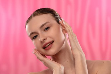 Açık tenli genç bir kadın nazikçe yüzüne dokunuyor, cilt teması ve gençlik ışıltısı öneriyor..