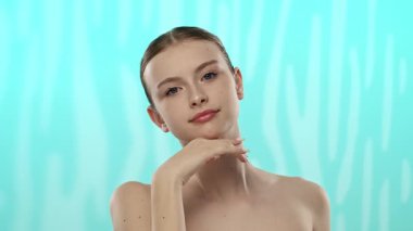 Yumuşak tenli ve topuz giymiş zarif saçlı genç bir kadın yumuşak bir turkuaz arka planda poz veriyor..