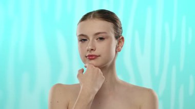 Yumuşak tenli ve topuz giymiş zarif saçlı genç bir kadın yumuşak bir turkuaz arka planda poz veriyor..