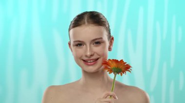 Bir kadın, canlı bir turuncu çiçeği gözünün üzerinde tutar. Yumuşak mavi desenli bir arkaplan, çarpıcı bir görsel karşıtlık yaratır..