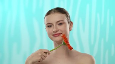 Nazik bir gülüşü olan genç bir kadın dingin mavi arka planda canlı bir turuncu gerbera çiçeği tutuyor..