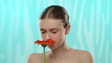 Yüzünde dingin bir ifade olan genç bir kadın parlak bir turuncu çiçeği yumuşak mavi arka planda tutuyor..