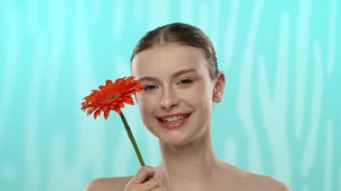 Nazik bir gülüşü olan genç bir kadın dingin mavi arka planda canlı bir turuncu gerbera çiçeği tutuyor..