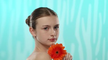 Nazik bir gülüşü olan genç bir kadın dingin mavi arka planda canlı bir turuncu gerbera çiçeği tutuyor..