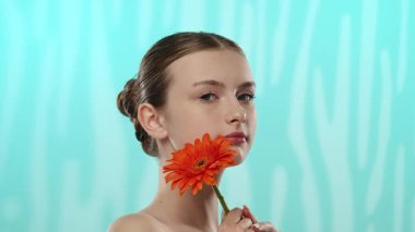 Nazik bir gülüşü olan genç bir kadın dingin mavi arka planda canlı bir turuncu gerbera çiçeği tutuyor..