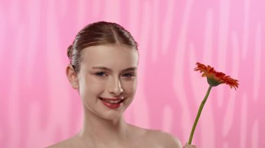 Genç bir kadın parlak turuncu bir gerbera papatyasını gözünün önünde canlı pembe bir arka plana karşı tutuyor..