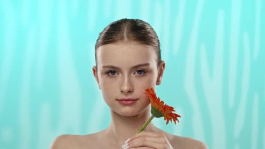 Nazik bir gülüşü olan genç bir kadın dingin mavi arka planda canlı bir turuncu gerbera çiçeği tutuyor..