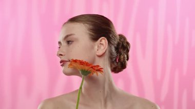 Genç bir kadın parlak turuncu bir gerbera papatyasını gözünün önünde canlı pembe bir arka plana karşı tutuyor..