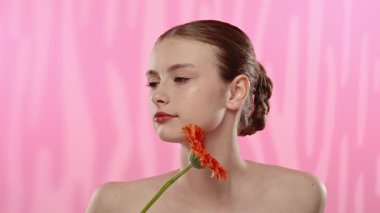 Genç bir kadın parlak turuncu bir gerbera papatyasını gözünün önünde canlı pembe bir arka plana karşı tutuyor..