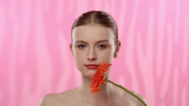 Genç bir kadın parlak turuncu bir gerbera papatyasını gözünün önünde canlı pembe bir arka plana karşı tutuyor..
