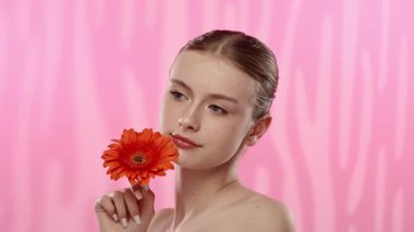 Genç bir kadın parlak turuncu bir gerbera papatyasını gözünün önünde canlı pembe bir arka plana karşı tutuyor..