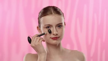 Genç bir kadın, sakin bir spa ortamında yüzüne yüz silindiri sürüyor. Yumuşak pembe bir arka planı var. Cilt bakımını teşvik ediyor..